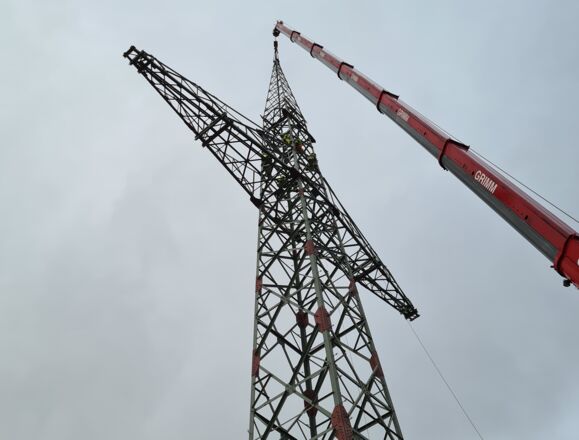 Grue GRIMM en action pour la construction d'une tour &eacute;lectrique haute tension avec des techniciens travaillant en hauteur.